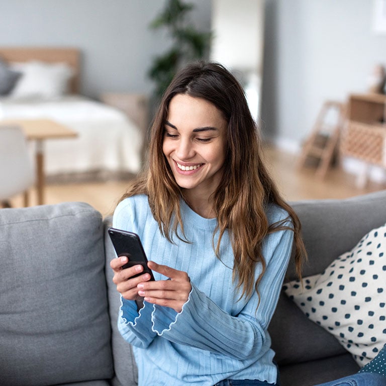 Cheerful-young-woman-using-mobile-phone-while-sitting-on-a-couch-at-home