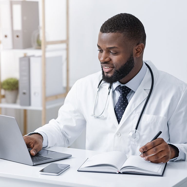 A doctor writes in a notebook while looking at a laptop