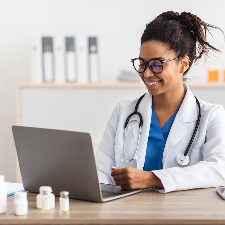A smiling doctor looks at her laptop screen sitting at a desk