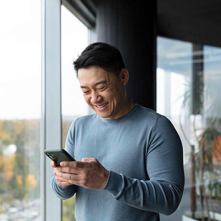 Smiling Asian man standing by a window and using a smartphone
